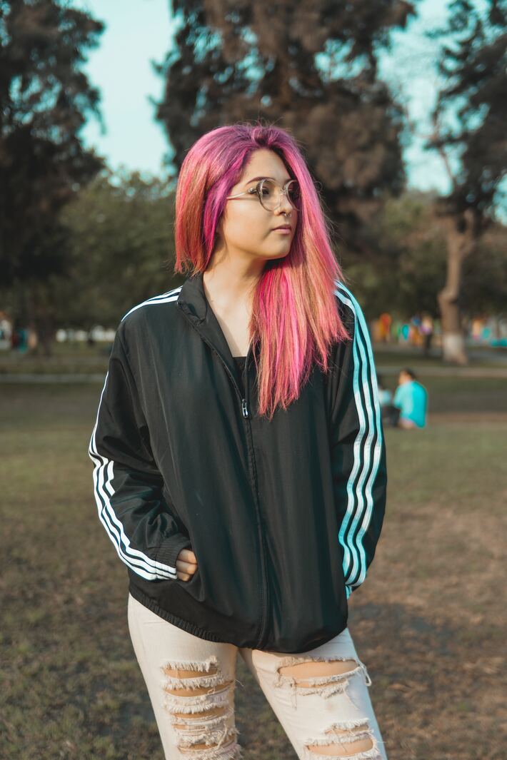 A young woman with glasses and bright pink and orange hair stands in a park looking into the distance